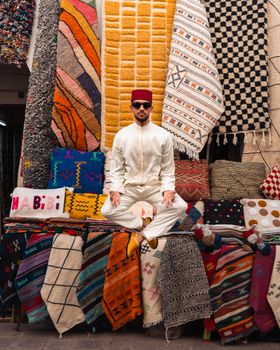 Man in traditional Moroccan clothing sits among colorful rugs in a market setting. Captivating blend of culture and textiles.