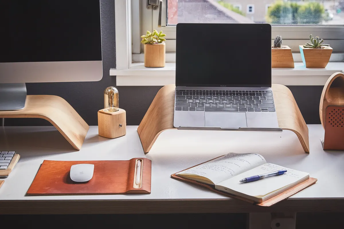 A switched-off laptop on a wooden stand at a tidy desk, with a notebook, a pen and potted plants by the window.