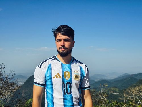 Free A man in an Argentina football jersey stands with a scenic mountain backdrop under a clear sky. Stock Photo