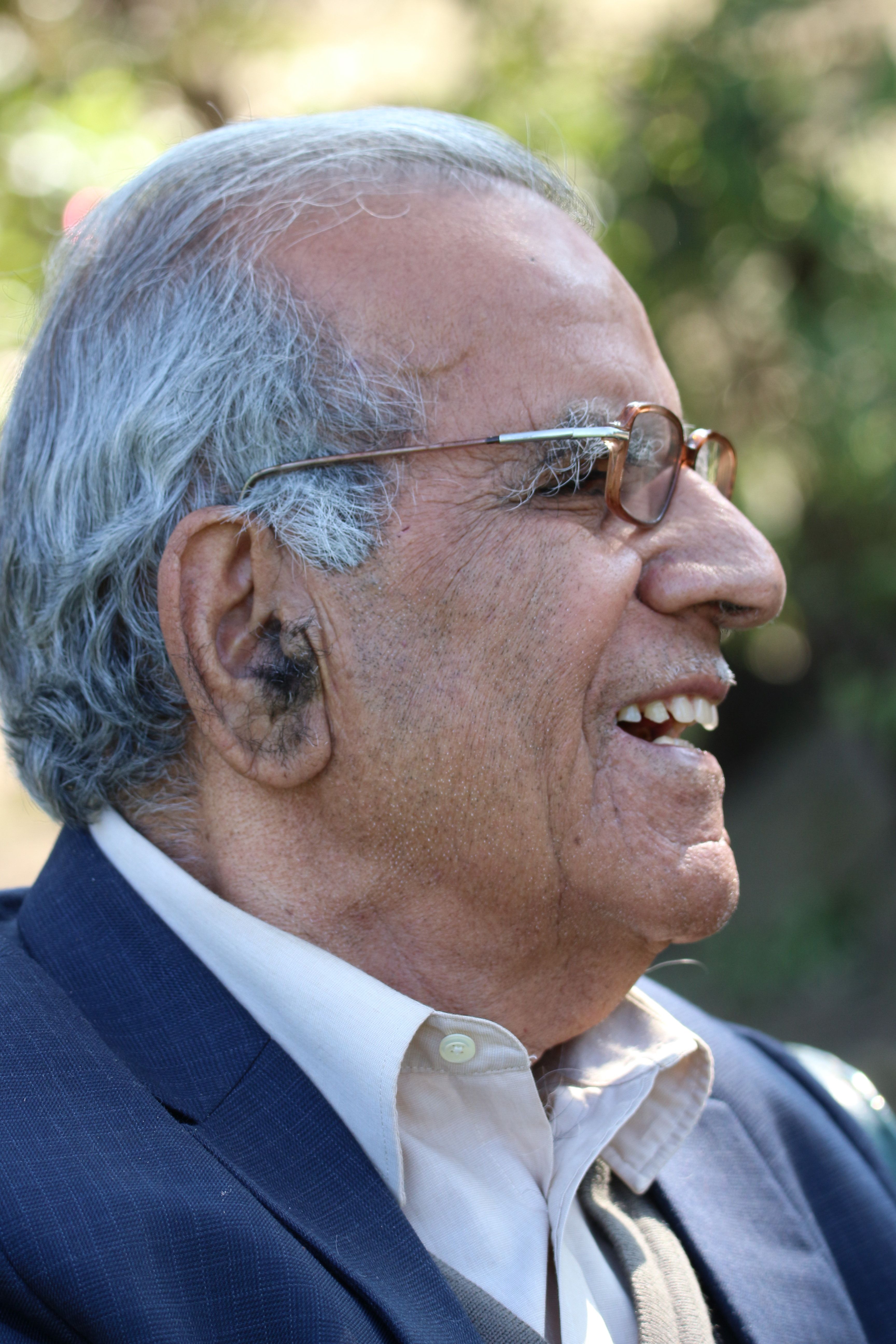 Profile of a smiling elderly man wearing glasses and a suit outdoors.