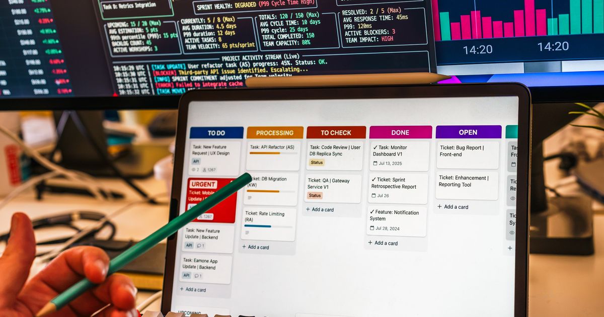A hand holding a pencil toward a tablet showing a task board, in front of monitors with charts.