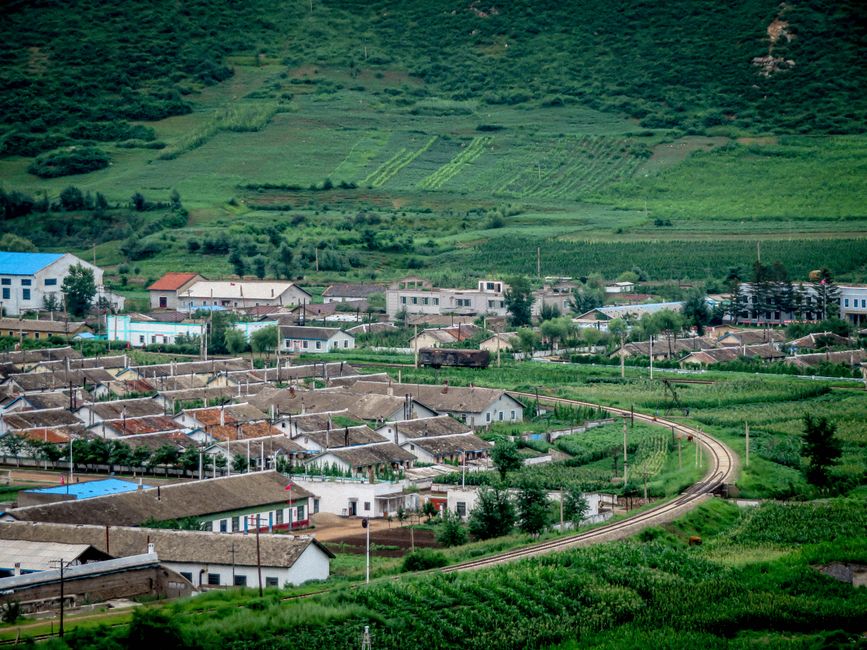 Lush green village landscape with railway curve amidst fields, showcasing rural architecture.
