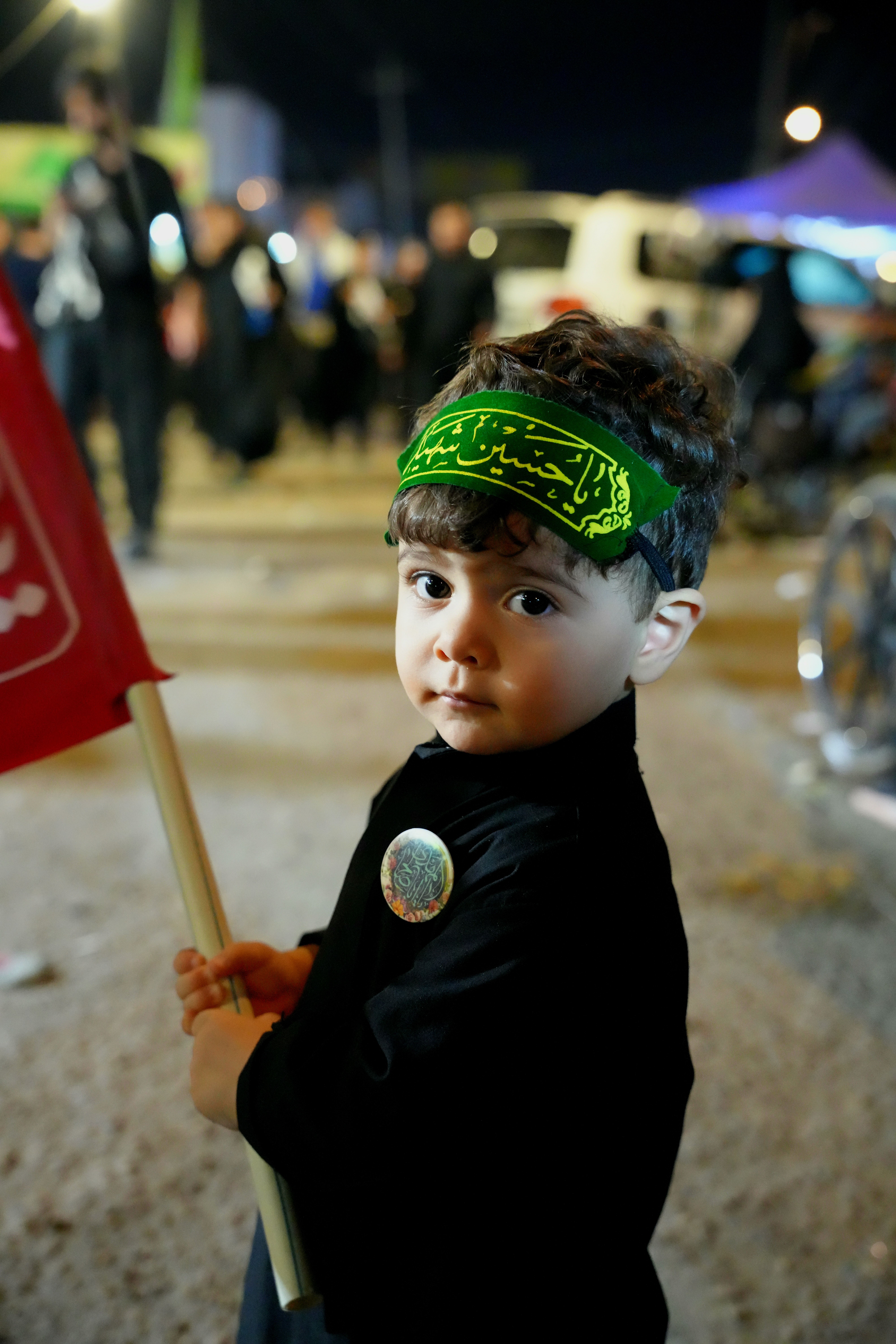 A young boy participates in the Arbaeen pilgrimage in Karbala, Iraq, holding a flag and wearing traditional attire.