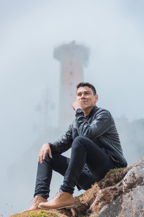 Man in Black Leather Jacket and Black Denim Jeans Sitting on Rock