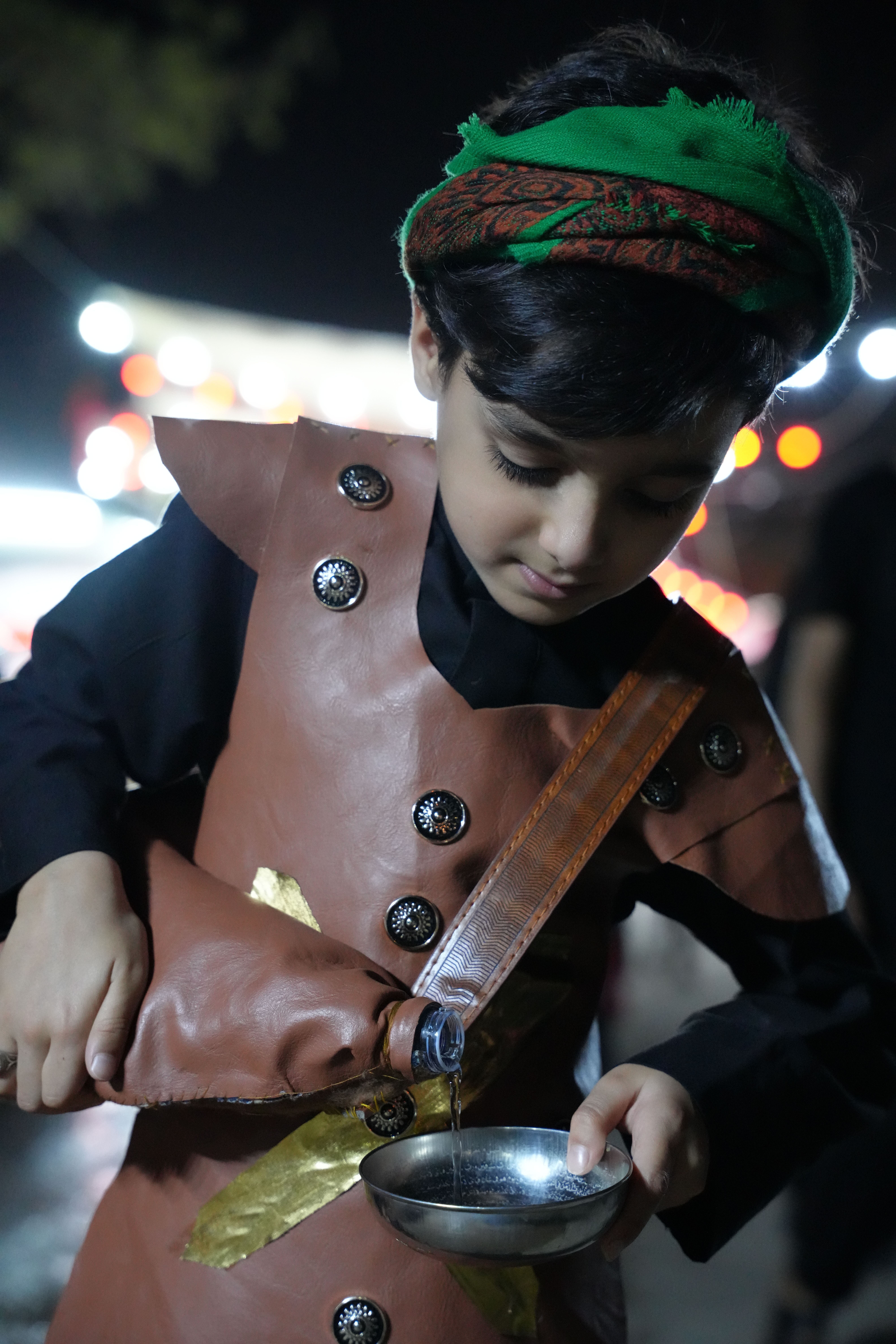 A young boy in traditional attire pouring water during Arbaeen pilgrimage in Karbala at night.