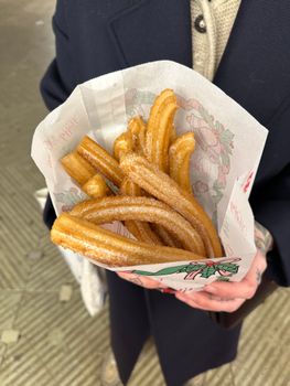 Crispy sweet churros in paper cone, held by person wearing coat, outdoor setting in Paris.