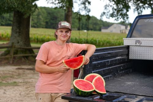 Free Smiling young farmer in a field holding a watermelon slice on a truck bed. Stock Photo