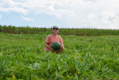 Free A smiling young farmer sits in a lush green field holding a watermelon, under a bright sky. Stock Photo