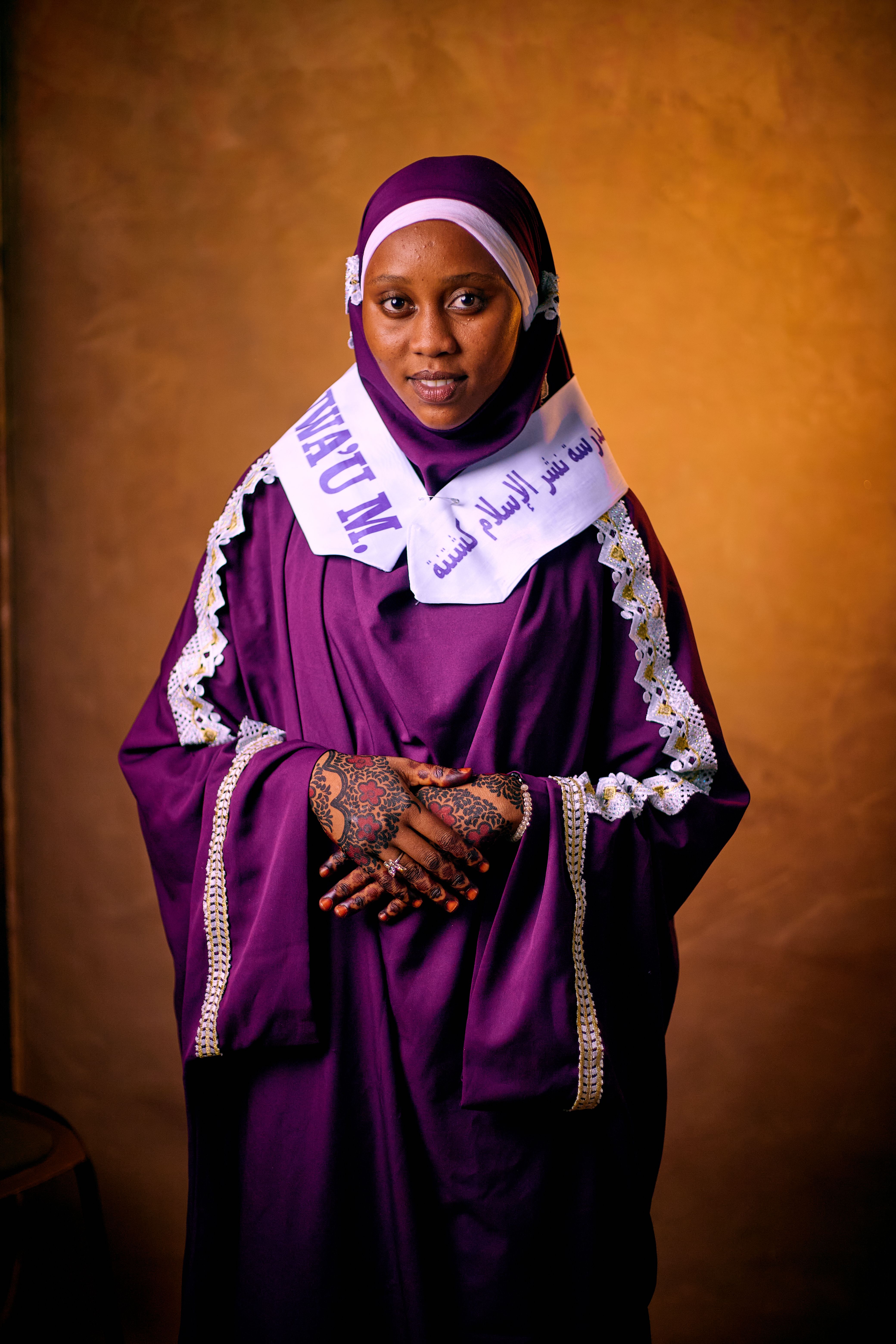 Woman in purple traditional dress with henna, standing against warm background.