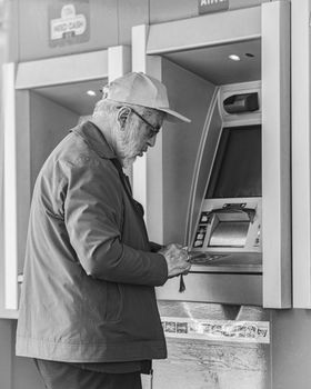 A senior man wearing a cap using an ATM. Black and white photograph.