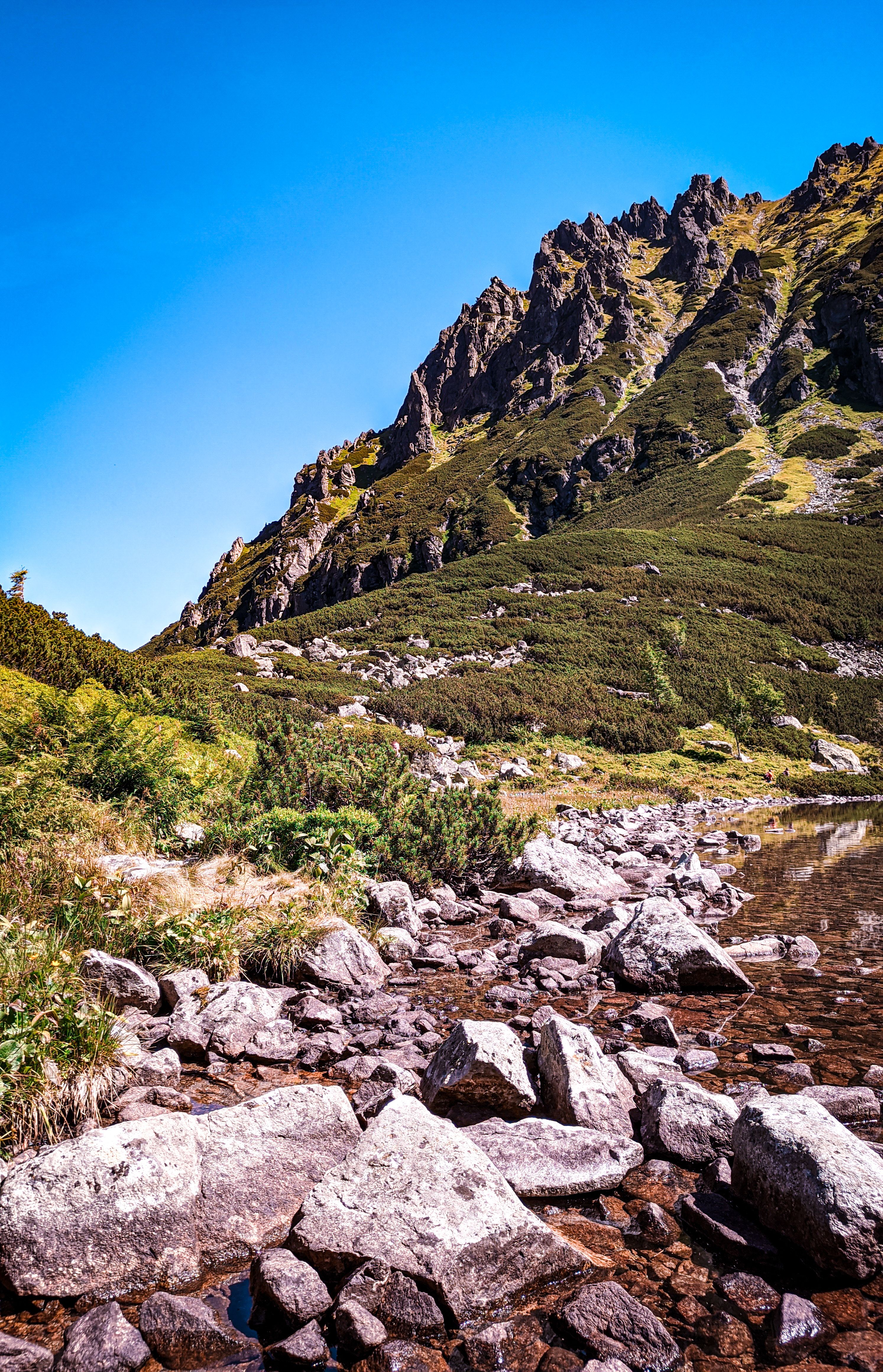 Majestic mountain landscape with a serene rocky lakeside under a bright blue sky.