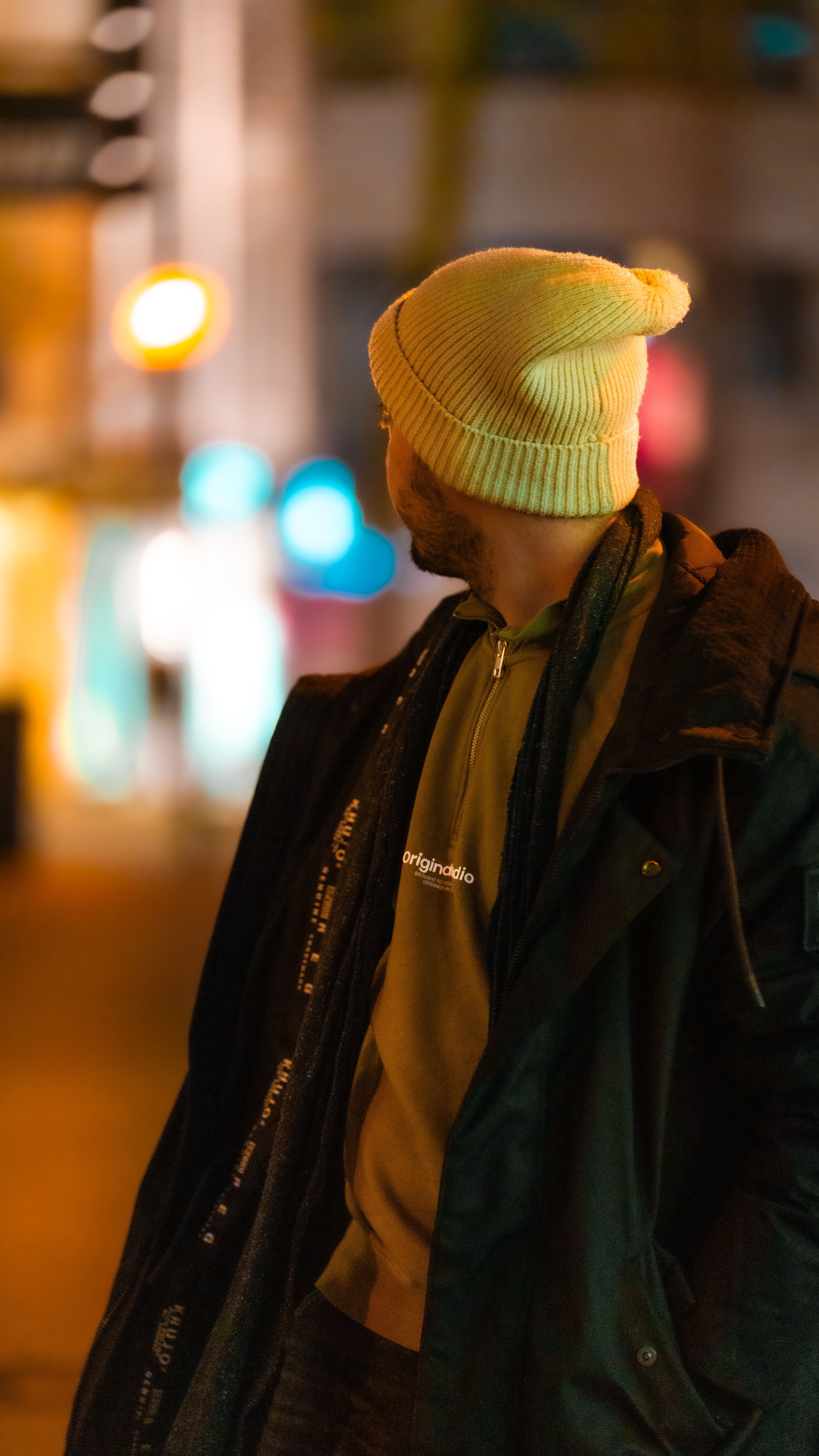A man in a beanie gazes at colorful urban lights on a vibrant night street.