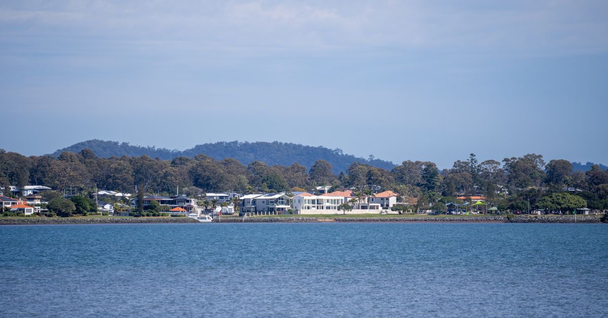 A tranquil view of a coastal residential area with lush trees and hills in the background under a clear blue sky.