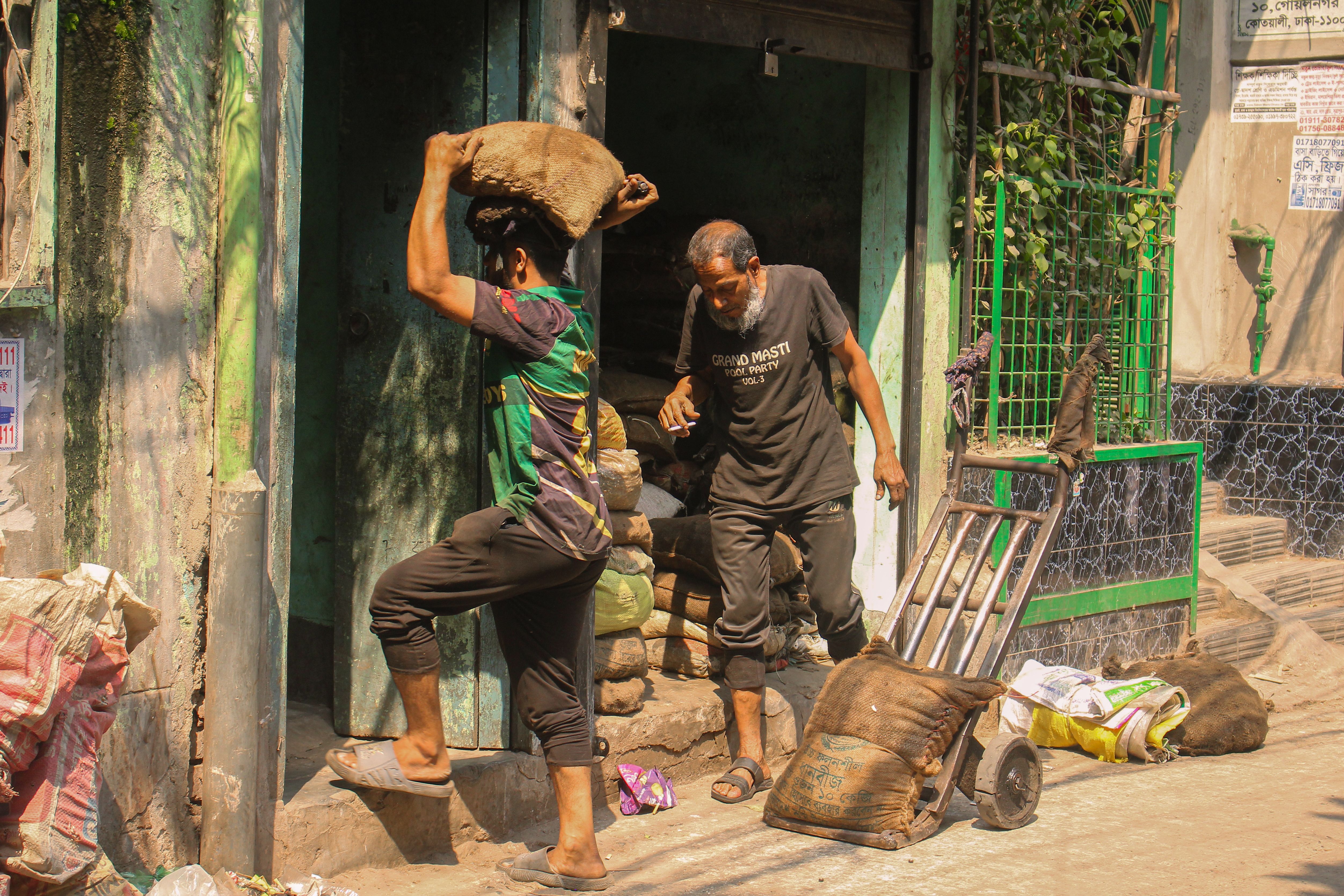 Two men working hard in a bustling street environment in Bangladesh, carrying heavy sacks outdoors.