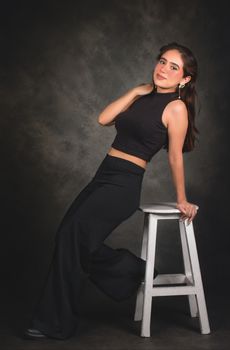 Young woman in black outfit poses elegantly on a stool indoors.