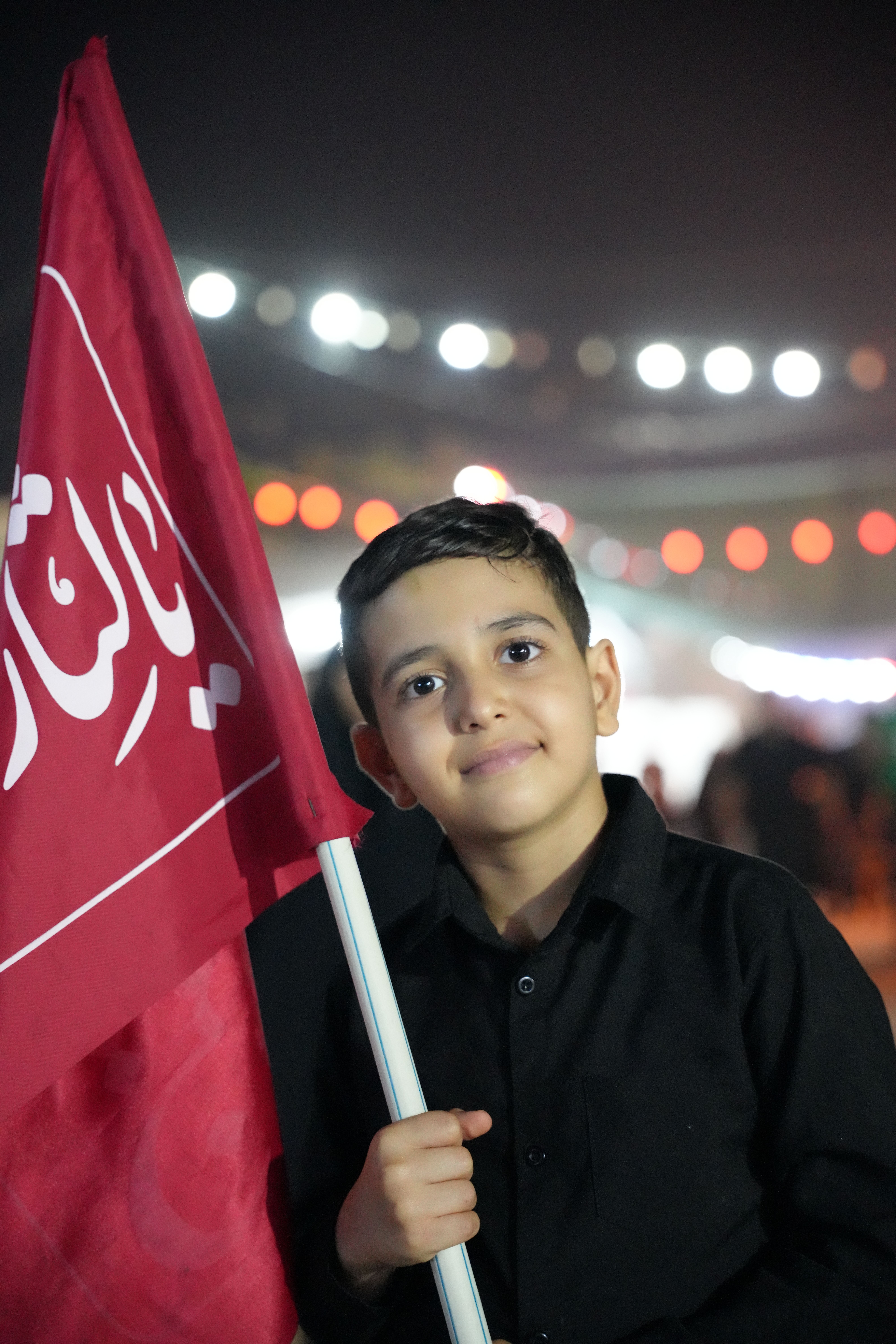 A young boy holds a red flag at night during Arbaeen in Karbala, Iraq, showcasing cultural and religious celebration.