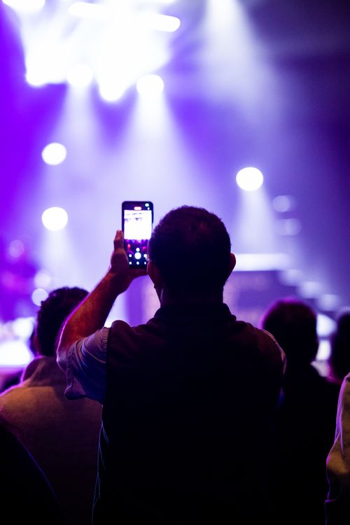 Free Audience member capturing a live concert on a smartphone, bathed in vibrant stage lighting. Stock Photo