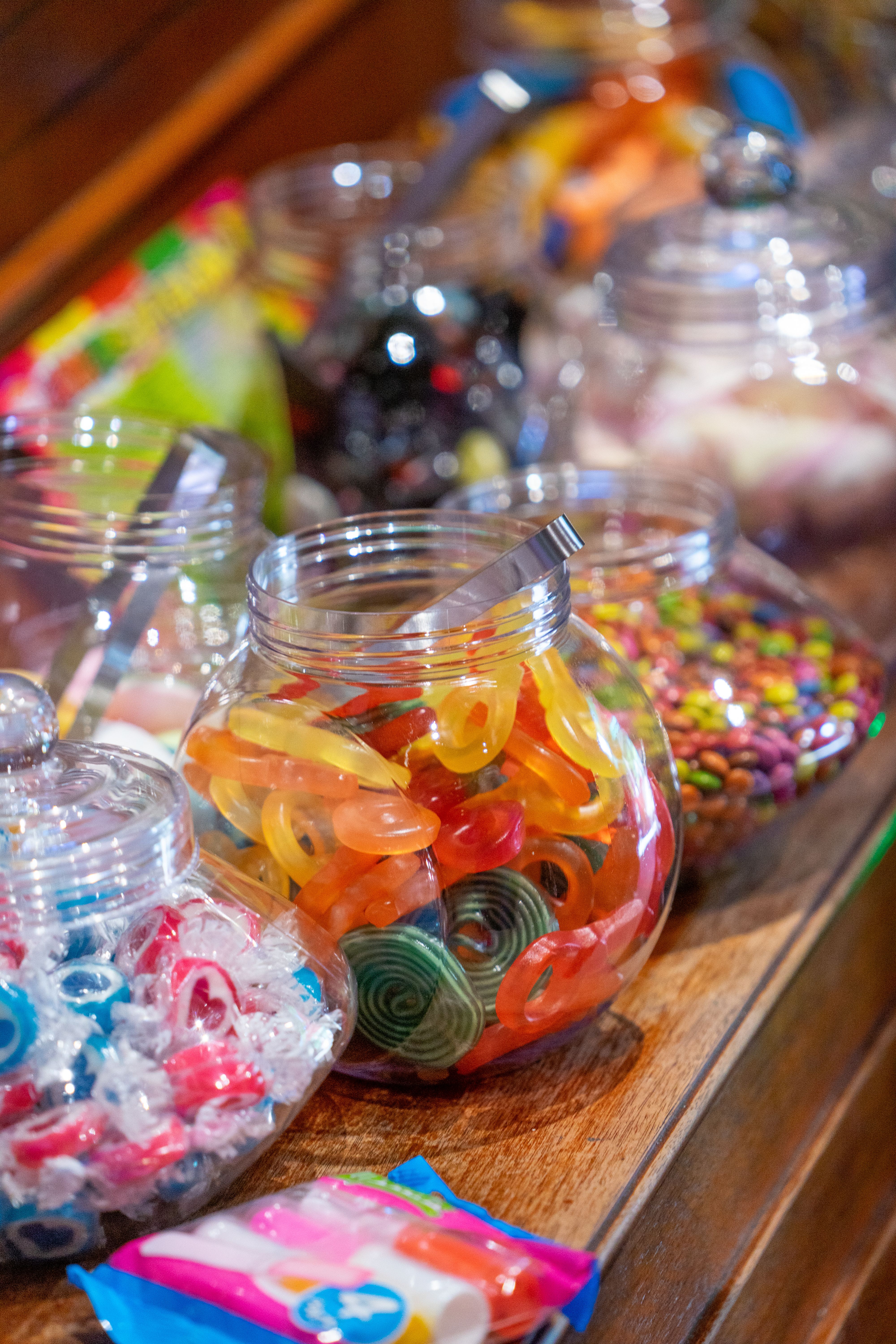 Bocaux en verre posés sur un comptoir de bois, remplis de bonbons gélifiés et de rouleaux de réglisse, une pelle en inox plantée dans l'un d'eux