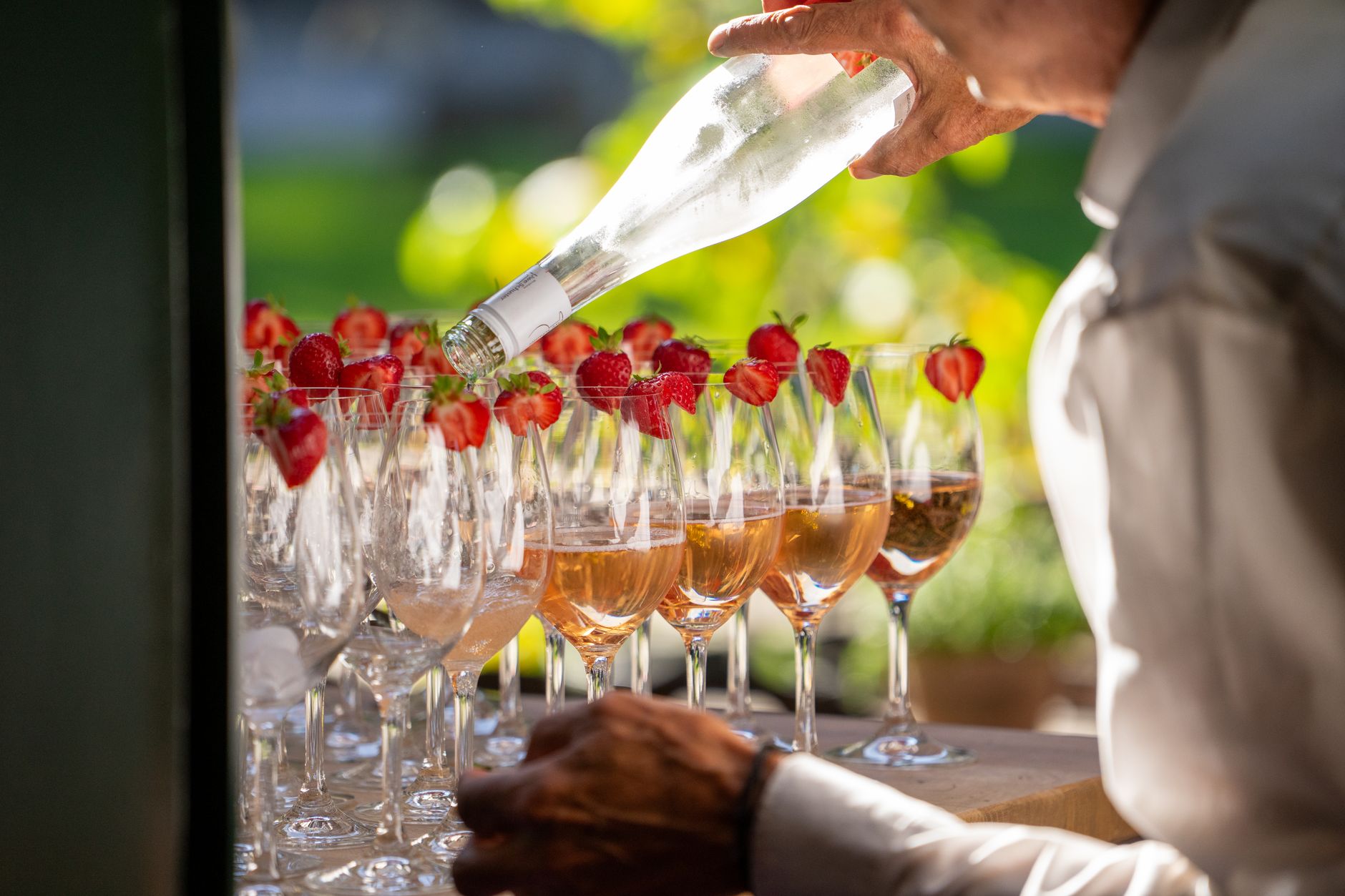 A person pours rosé wine into glasses adorned with strawberries at an outdoor party.