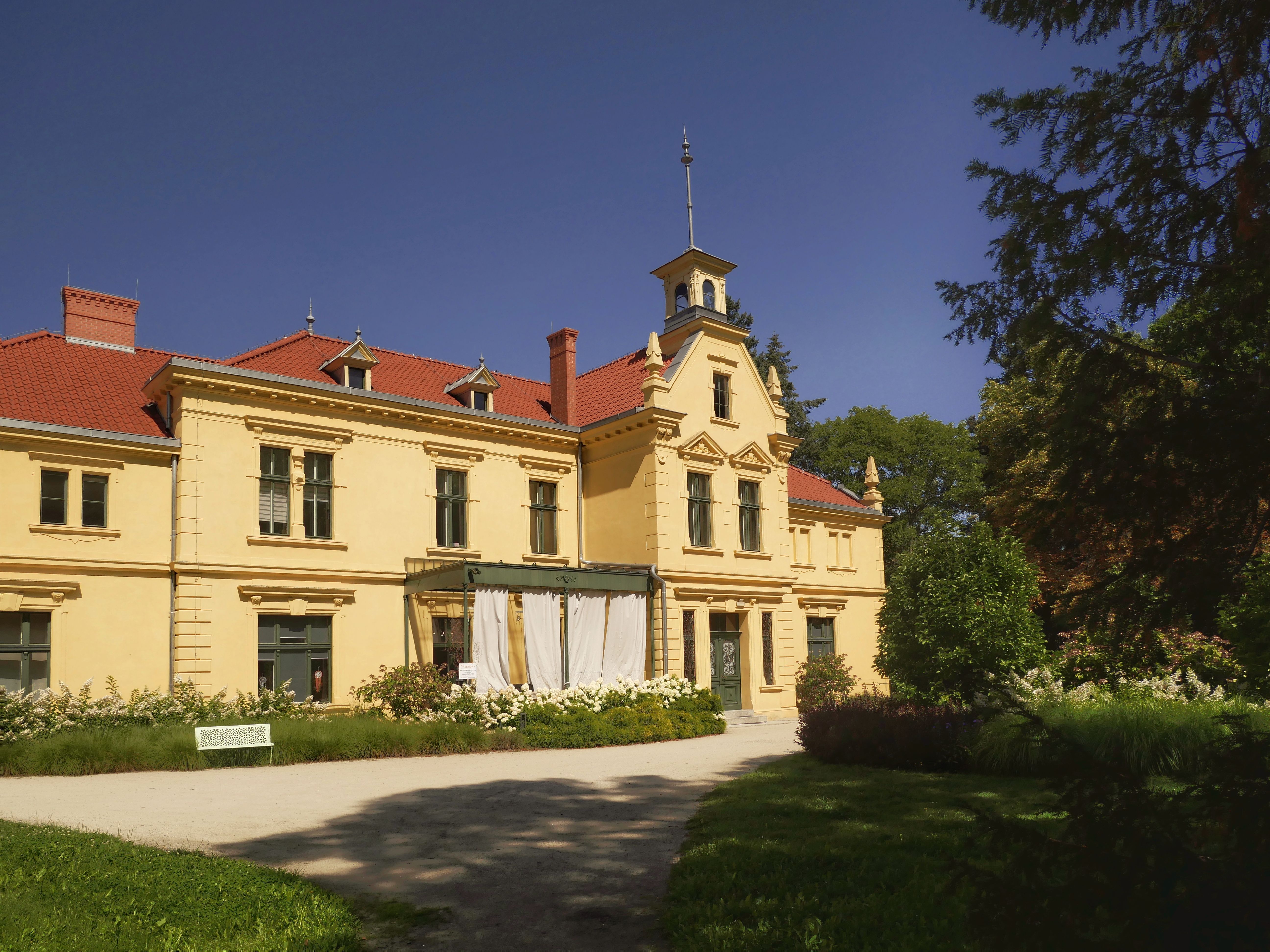 Elegant yellow manor house with red tile roof surrounded by lush gardens in Zielona Góra, Poland.