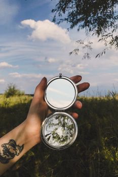 A hand with a tattoo holds an open compact mirror reflecting the sky and nature.