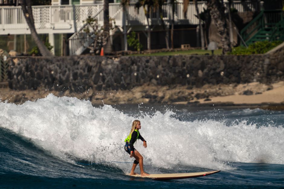 Blonde surfer girl navigating ocean waves near Hawaiian beach houses.