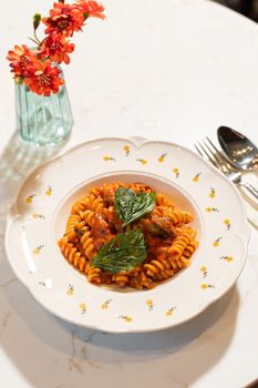 Delicious fusilli pasta with tomato sauce and fresh basil served in a floral ceramic bowl on a marble table.