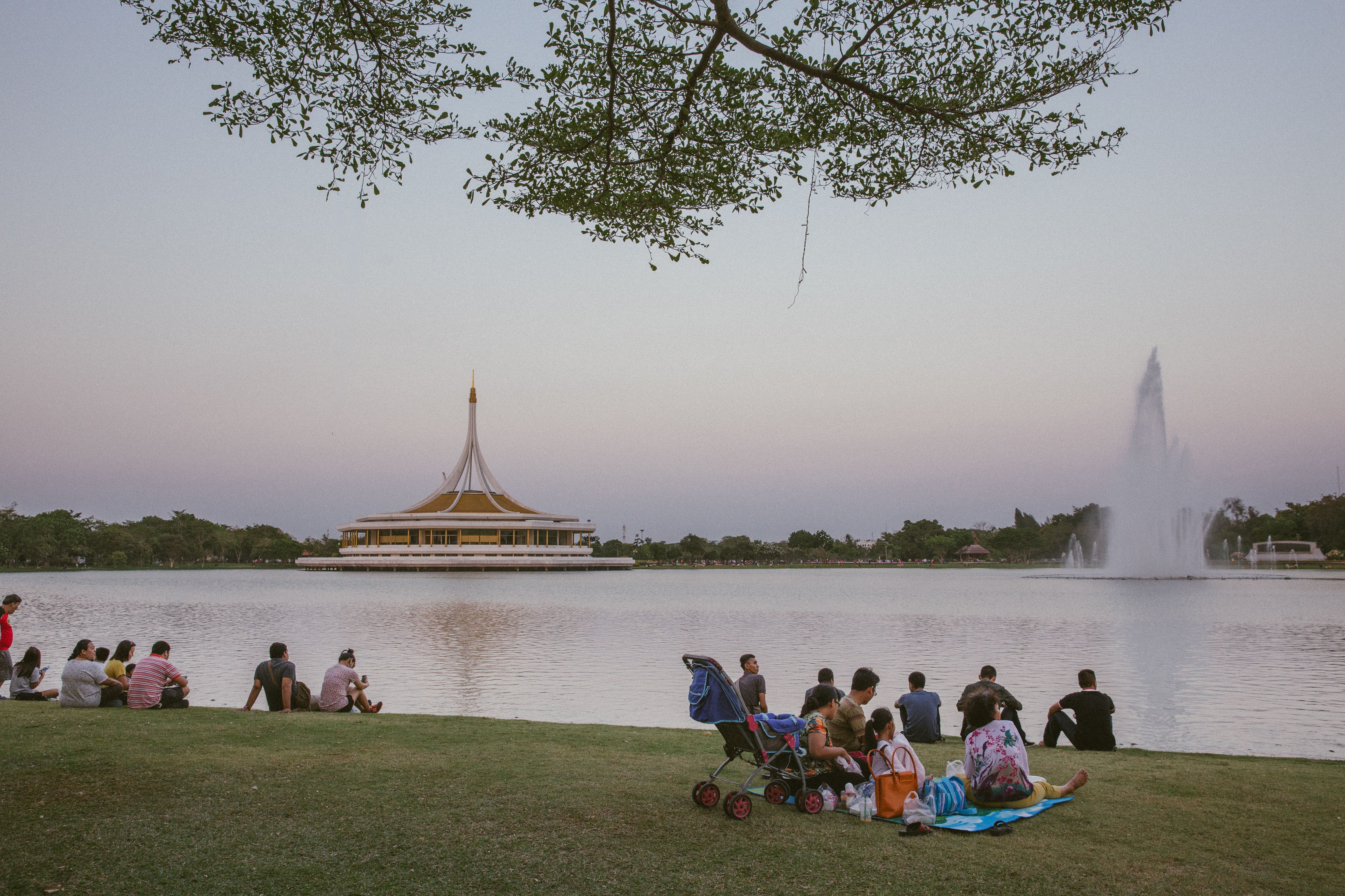 People relaxing by the lake at Suan Luang Rama IX Park in Bangkok during sunset with fountain and pavilion view.