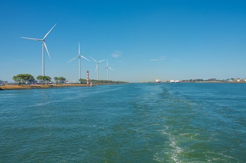 Free Wind turbines on a riverbank with a clear blue sky, showcasing renewable energy in a serene environment. Stock Photo