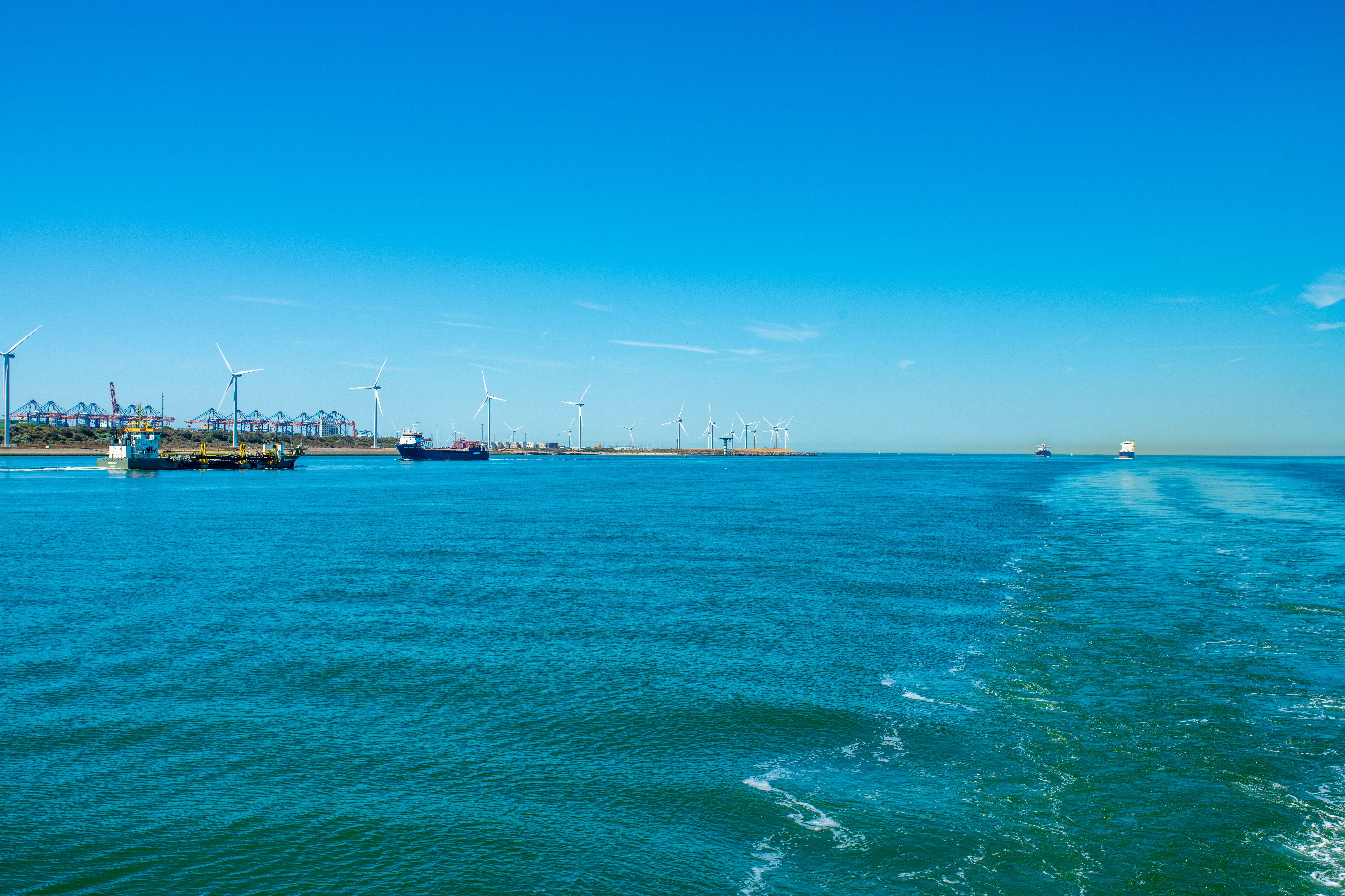 Temsilî fotoğraf: A scenic view of wind turbines and cargo ships across a vibrant blue sea under a clear sky.