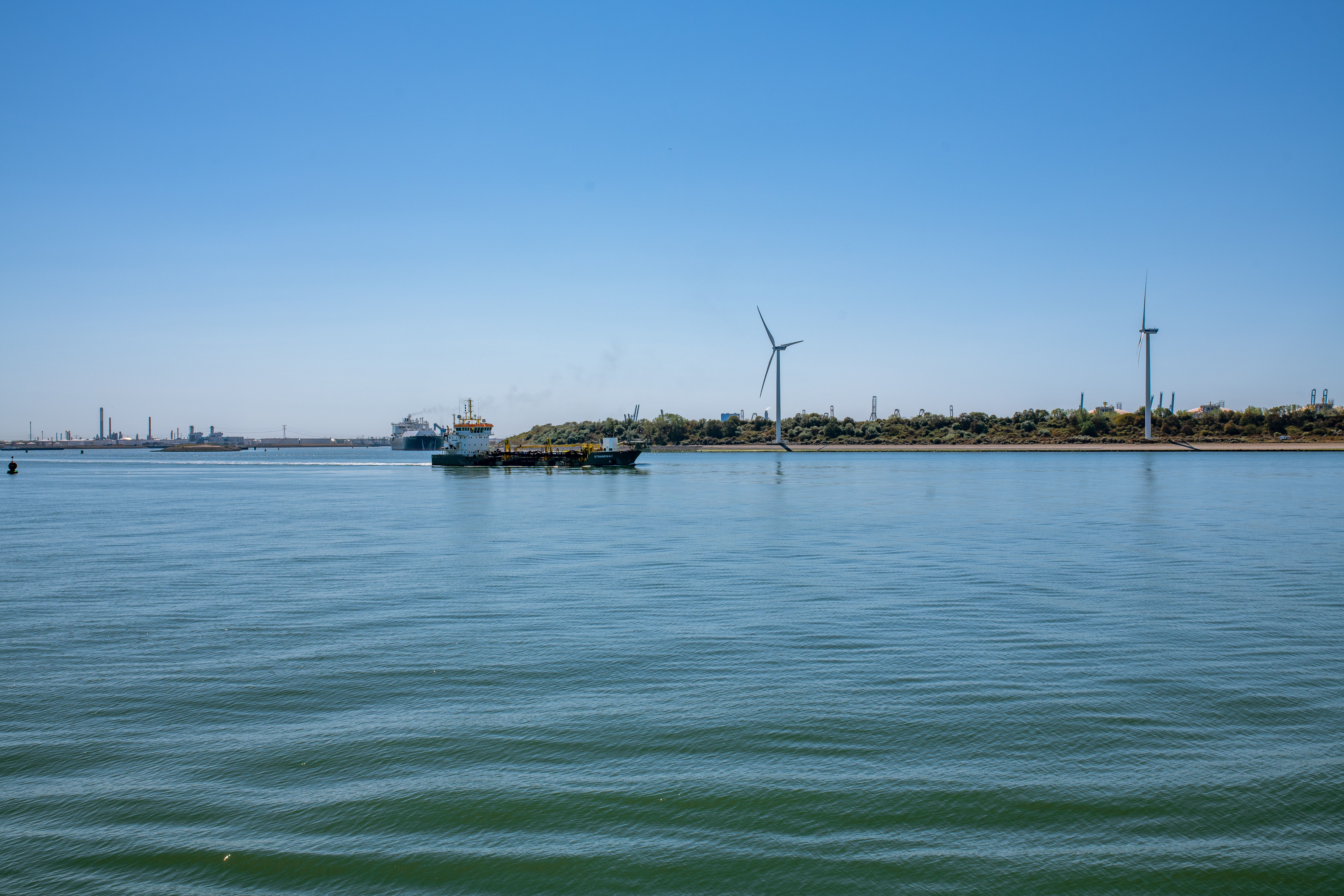 A cargo ship sails near wind turbines on a calm sea with clear blue skies.