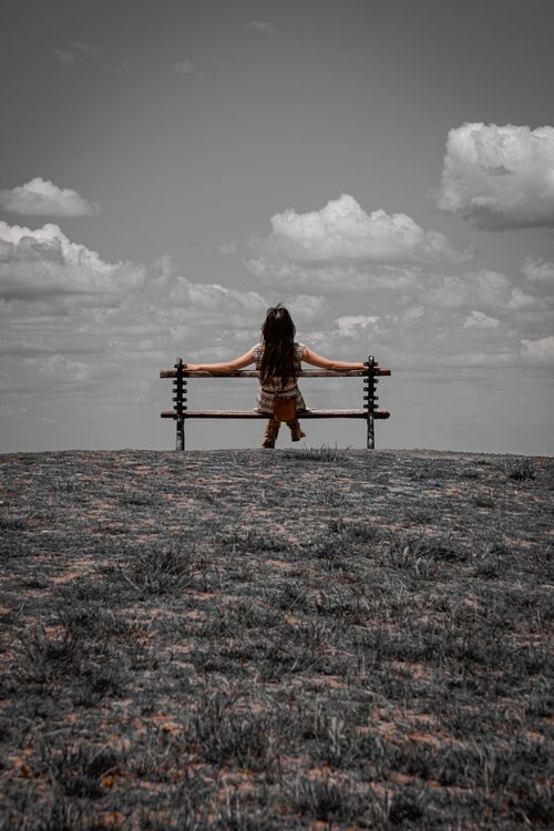 Free Girl sitting alone on a bench under dramatic clouds in an open field. Serene and thoughtful scene. Stock Photo
