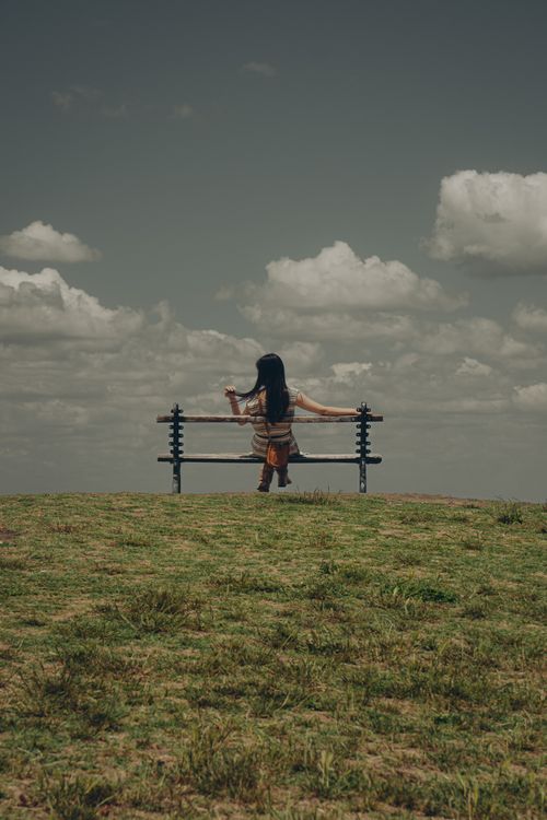 Free Woman enjoys tranquil view from a park bench under cloudy skies. Perfect for relaxation and travel themes. Stock Photo