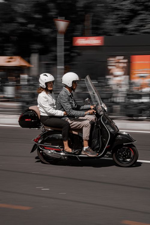 Free Man and woman on a scooter with helmets driving through city street. Stock Photo