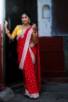A woman in a red sari and gold jewelry posing gracefully by a doorway, emphasizing cultural elegance.