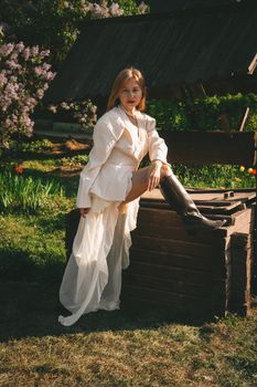 Stylish woman in elegant white ensemble poses outdoors by a wooden structure on a sunny day.