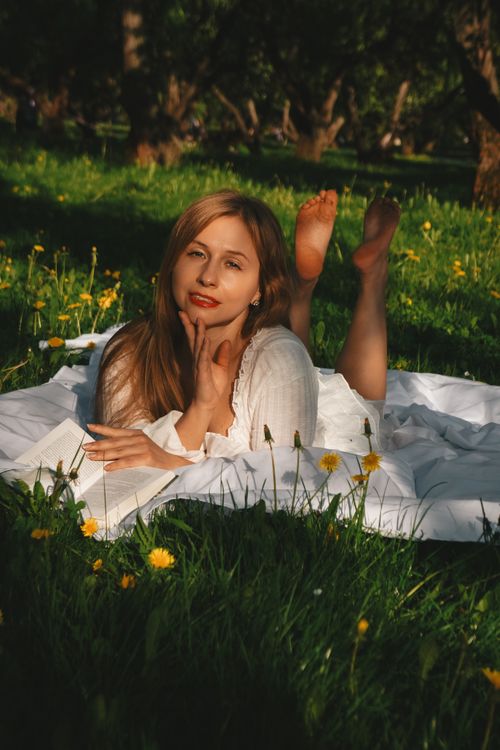 Free A young woman lying on a blanket in a sunny meadow, surrounded by dandelions, smiling and reading a book. Stock Photo