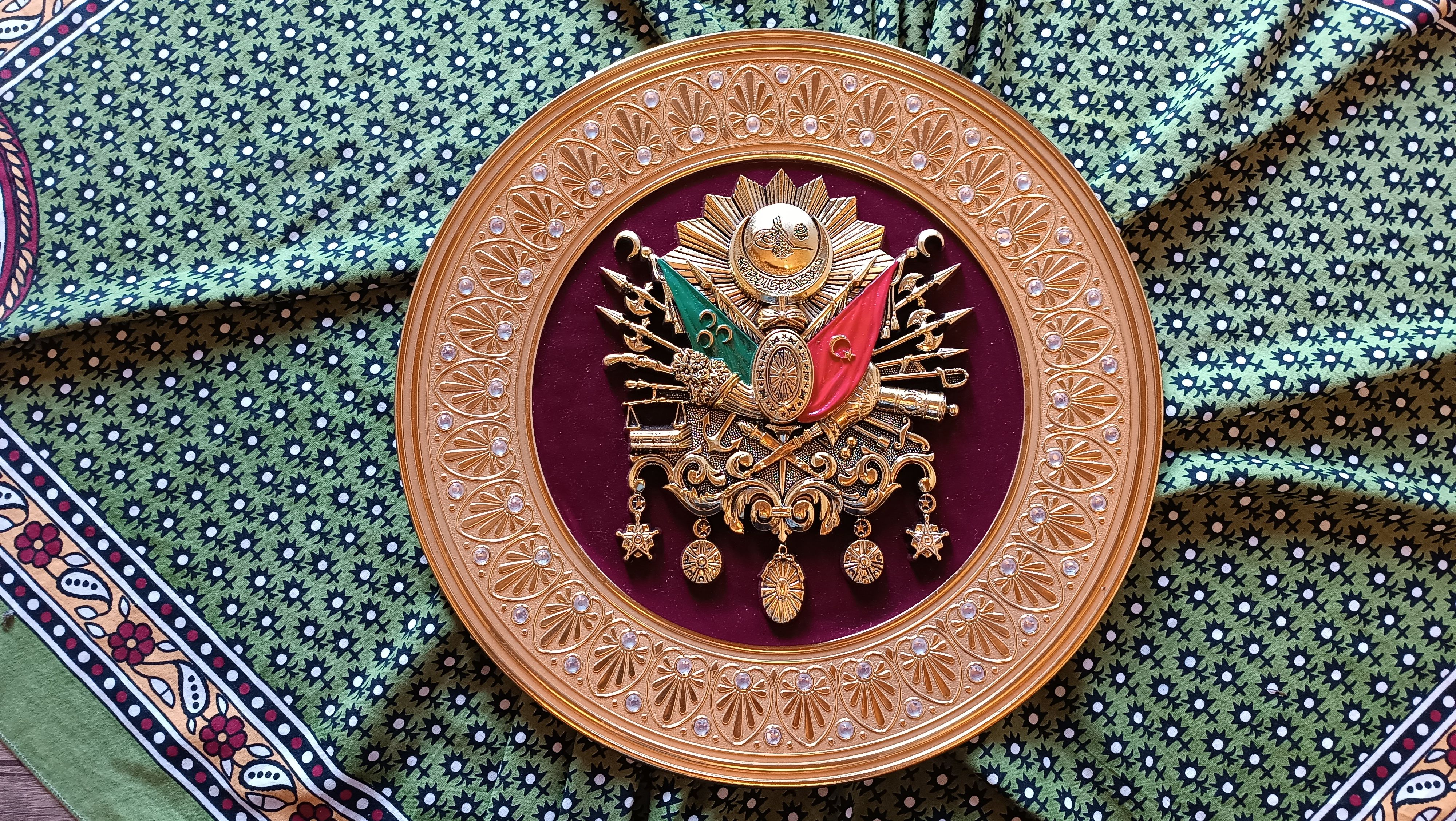 A detailed Ottoman coat of arms emblem with ornate detailing set against a patterned fabric background.