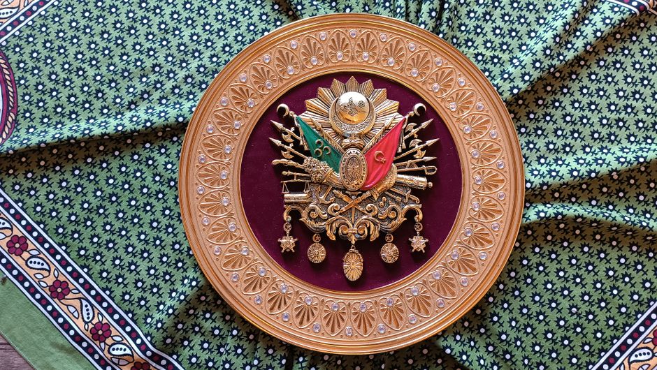 A detailed Ottoman coat of arms emblem with ornate detailing set against a patterned fabric background.