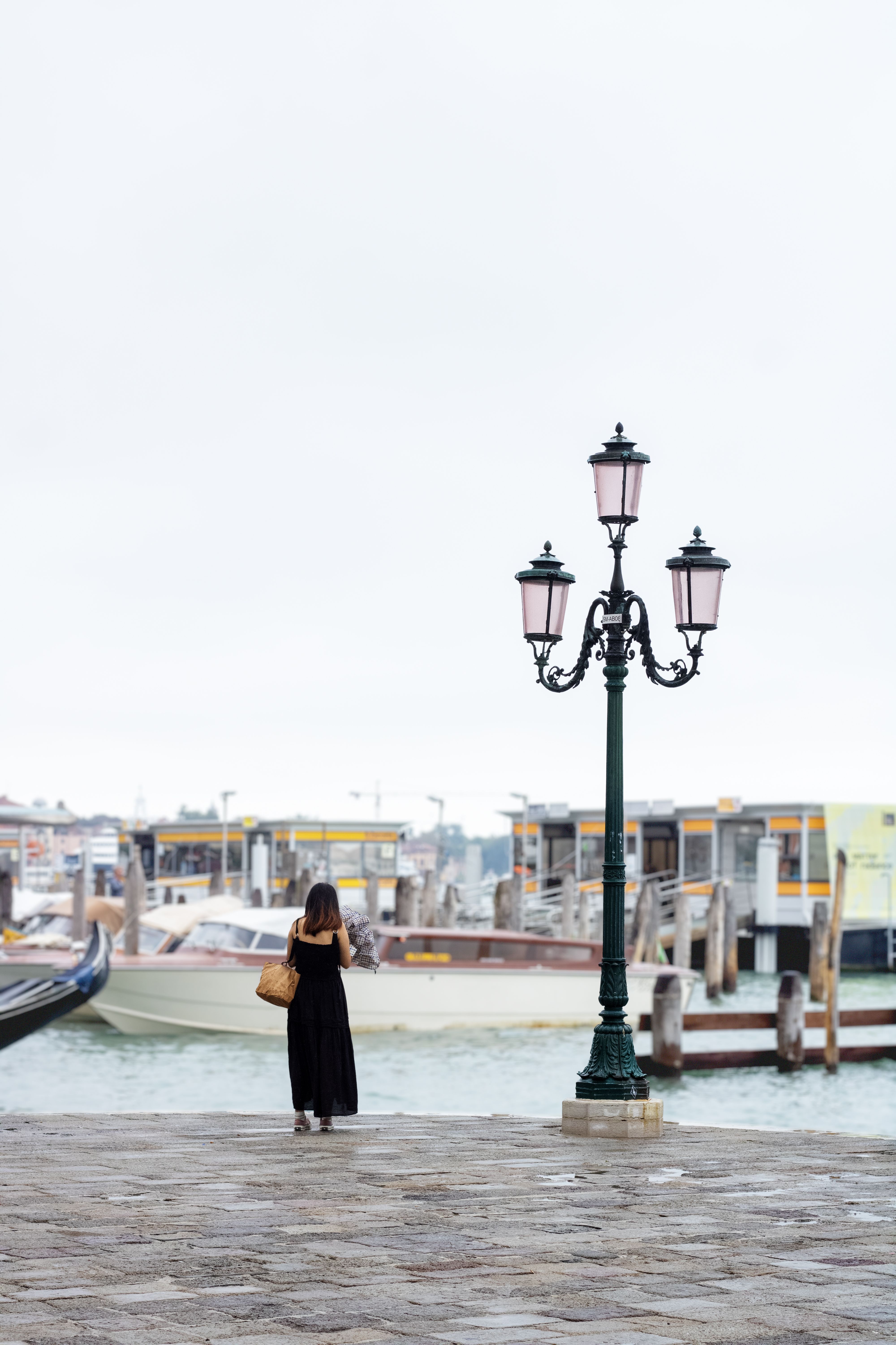 A woman stands alone by the Venetian waterfront under a classic lamp post, capturing the peaceful scene.