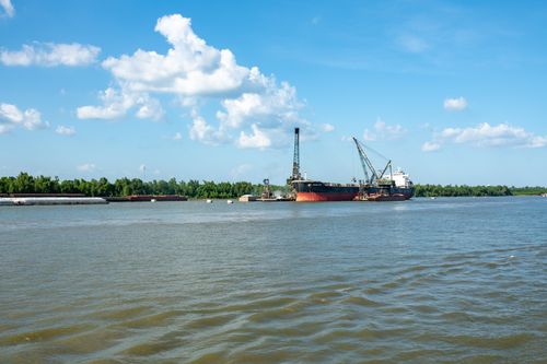 Free A cargo ship docked at a river port under a bright blue sky with fluffy clouds. Stock Photo