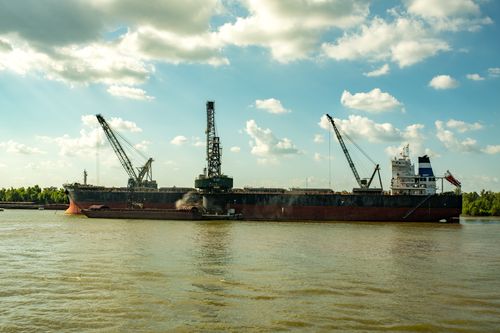 Free A large cargo ship equipped with cranes sailing on a tranquil river against a bright sky. Stock Photo