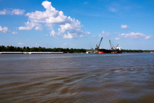 Free A cargo ship with cranes navigates a wide river under a bright blue sky filled with fluffy clouds. Stock Photo