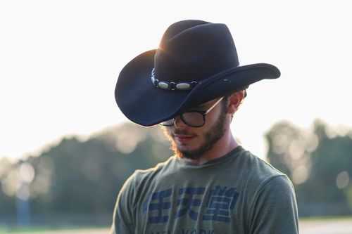 Free A young man wearing a cowboy hat and glasses is outdoors at sunset, conveying a relaxed, western style. Stock Photo