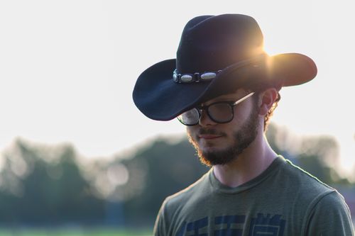 Free A young man with glasses wearing a cowboy hat in a sunlit outdoor setting. Stock Photo