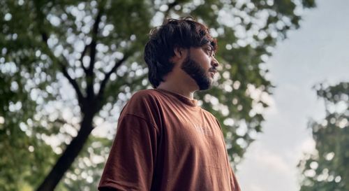 Free A man stands thoughtfully outdoors with lush trees in the background. Stock Photo