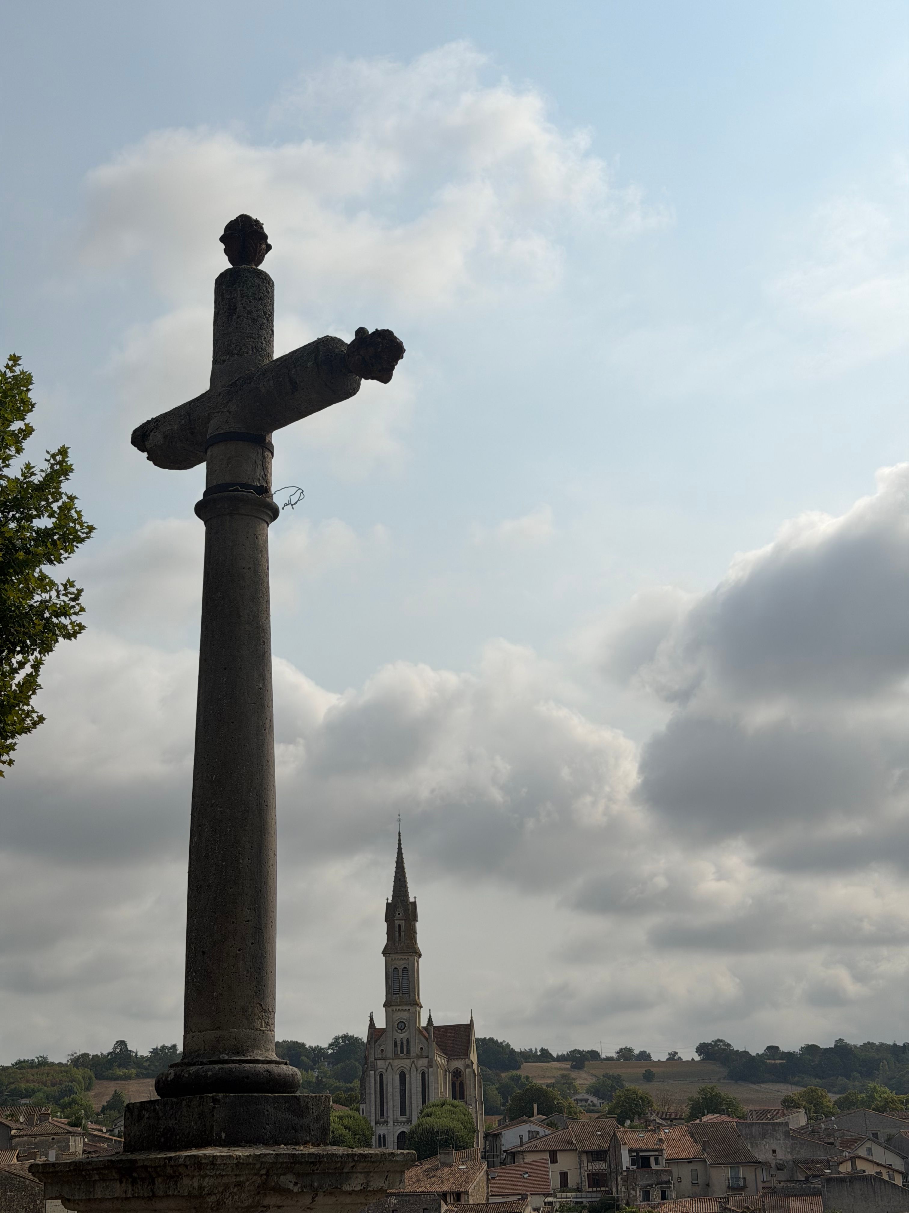A scenic view of a historic cross with a French village and church in the background under a cloudy sky.