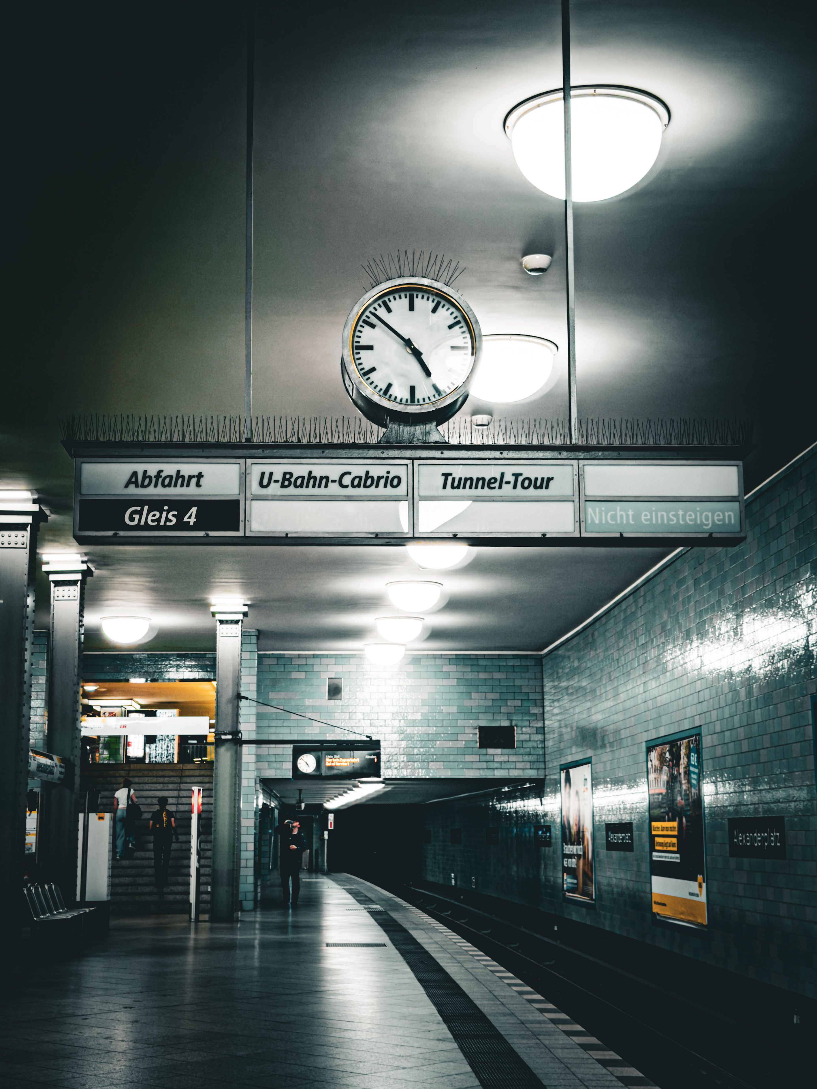 Empty Berlin subway station platform at night with a clock and signs overhead, creating a moody ambiance.