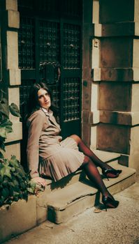 Stylish woman in vintage attire sits gracefully by a rustic doorway, evoking a classic mood.