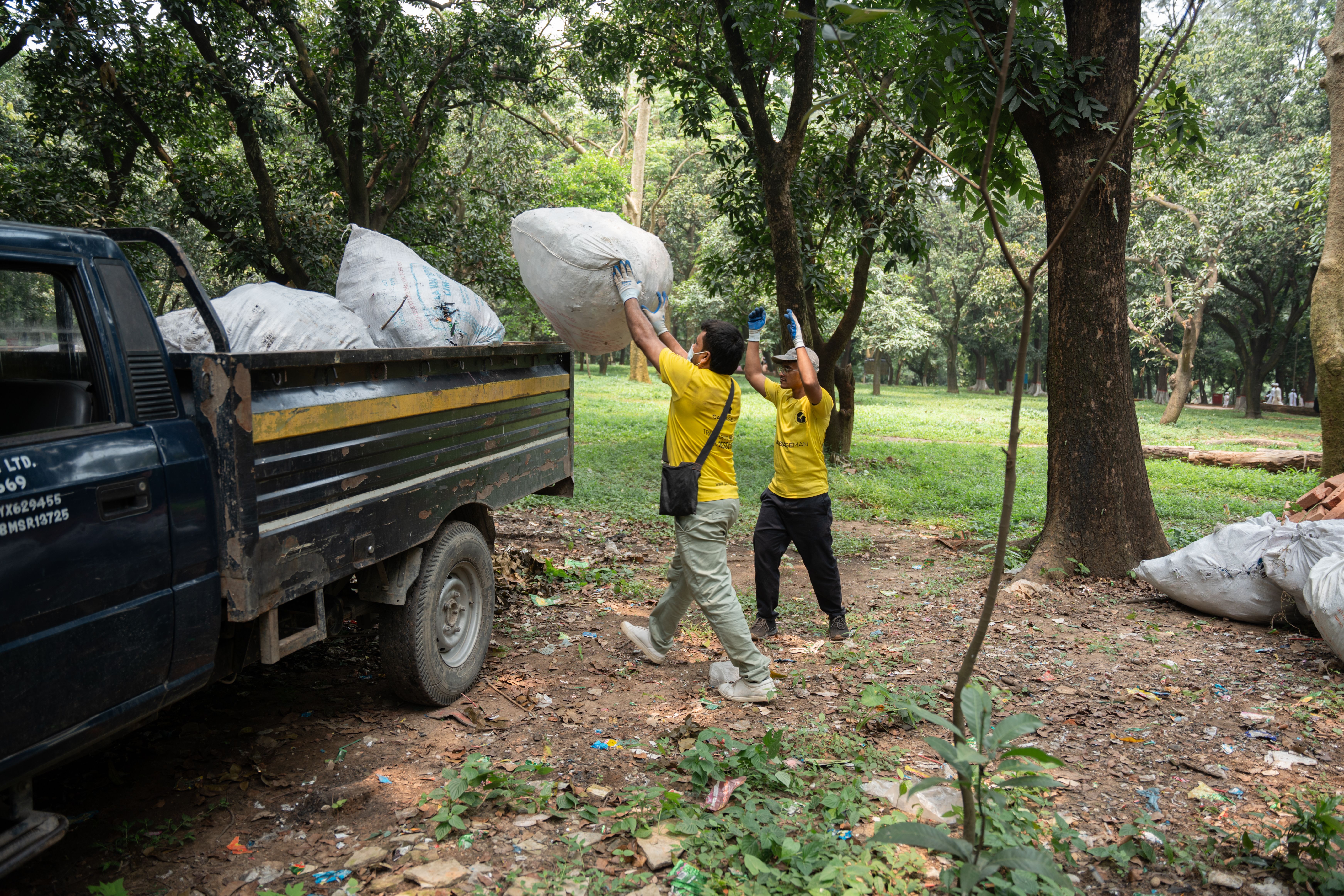 Two workers in yellow shirts load a truck with large garbage bags in a wooded park area